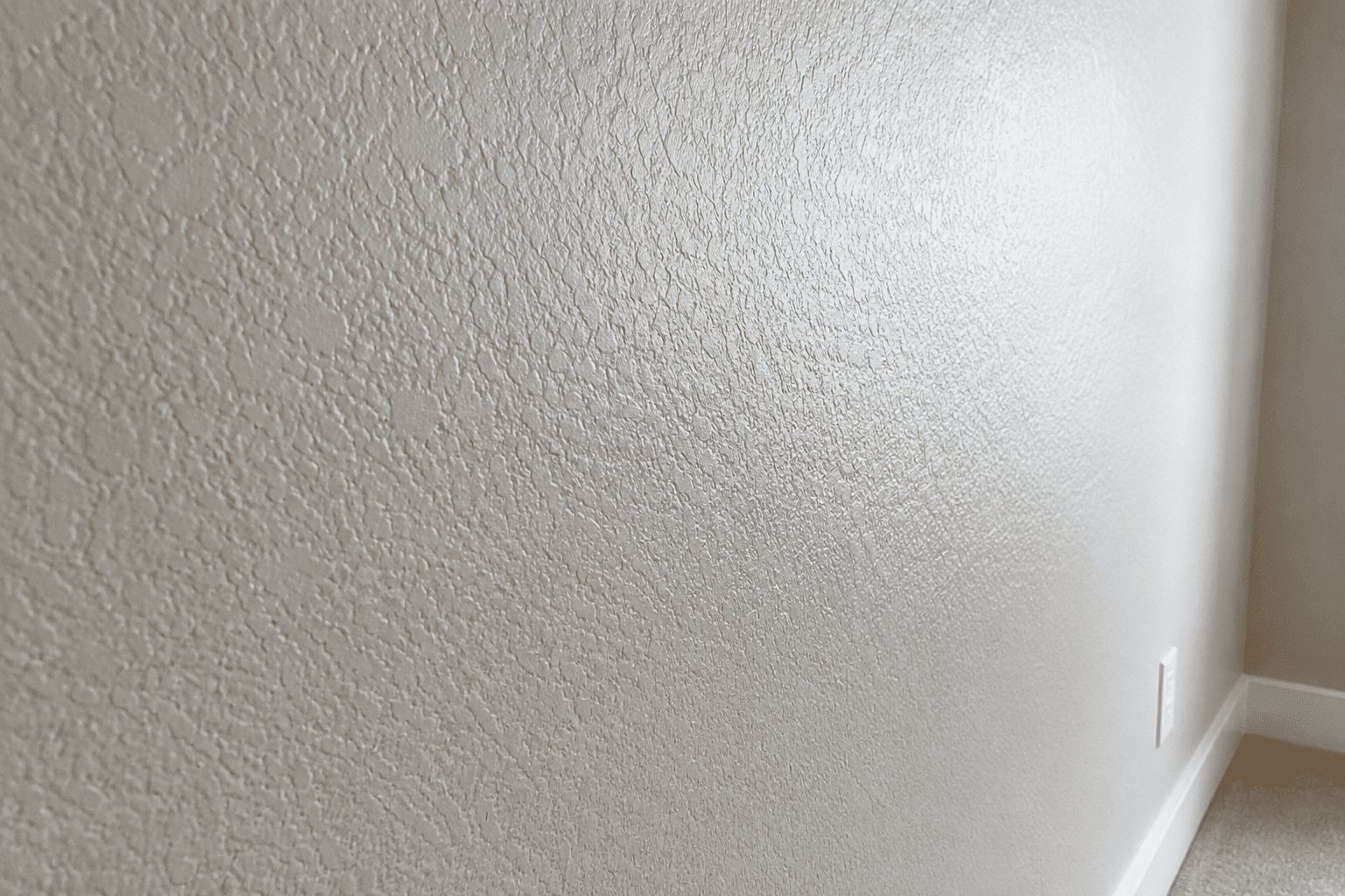 Light beige wall with a heavy orange peel texture, white baseboard, and carpeted floor.