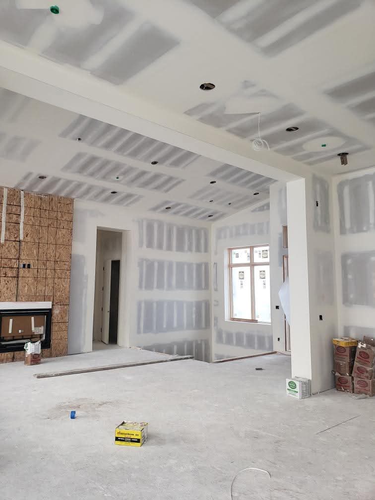 Interior room under construction with mudded drywall, recessed lighting holes, and unfinished concrete floors.