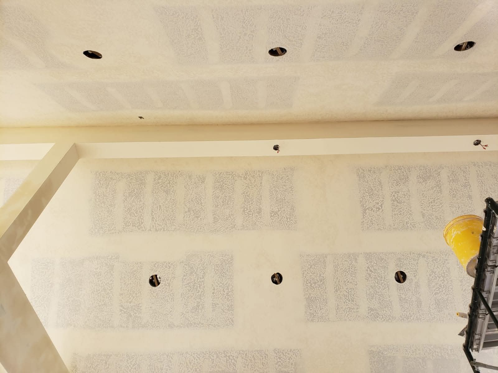 Drywall ceiling and wall with textured patches and circular cutouts for recessed light fixtures.